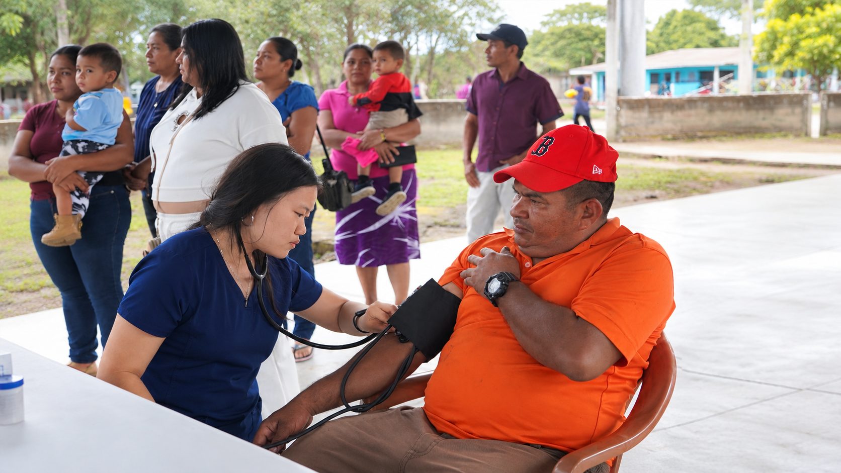 A Mercy Health Center clinician checking a patient's blood pressure at an outdoor community clinic while neighbors wait nearby