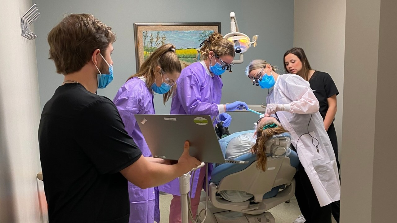 Mercy dental team members in purple gowns and blue masks treating a patient reclined in a dental chair while a colleague holds a laptop open beside them